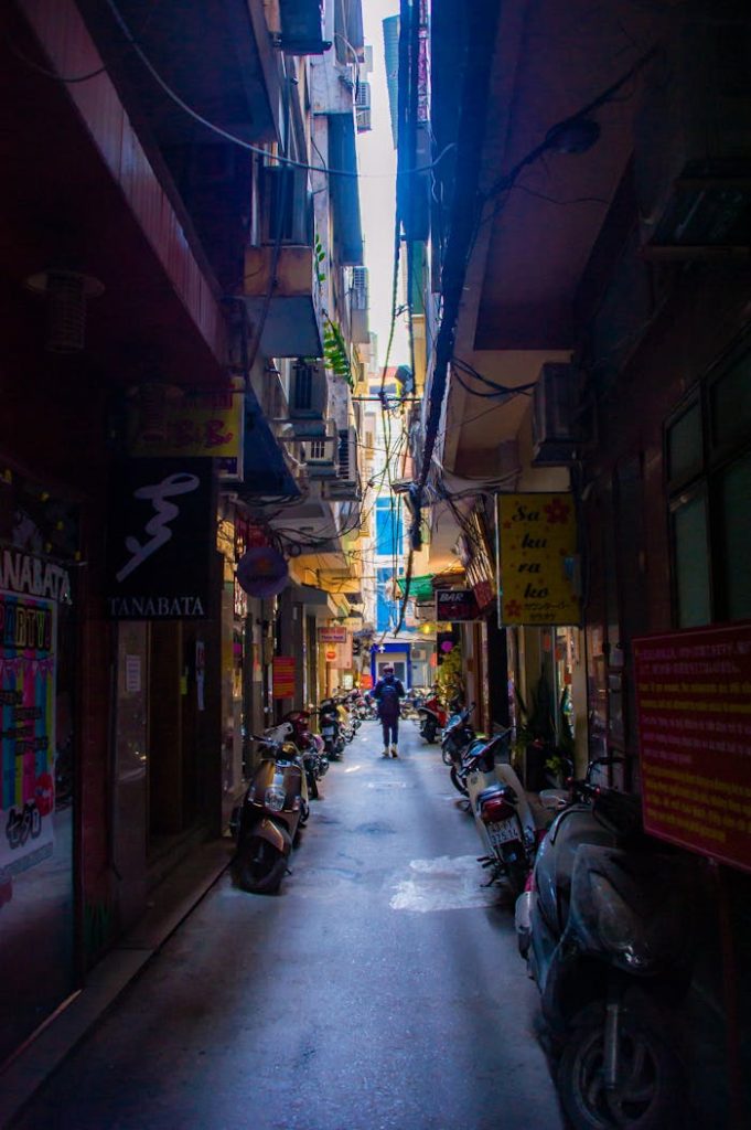 A bustling narrow alleyway filled with shops and scooters under daylight.